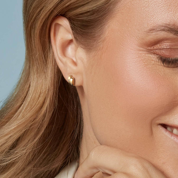 Close-up van een vrouw die glimlacht en haar oor laat zien versierd met Boccia Titanium Oorstekers 0552-06. Haar hand rust bij haar kin en haar lichtbruine haar is achter haar oor verborgen.