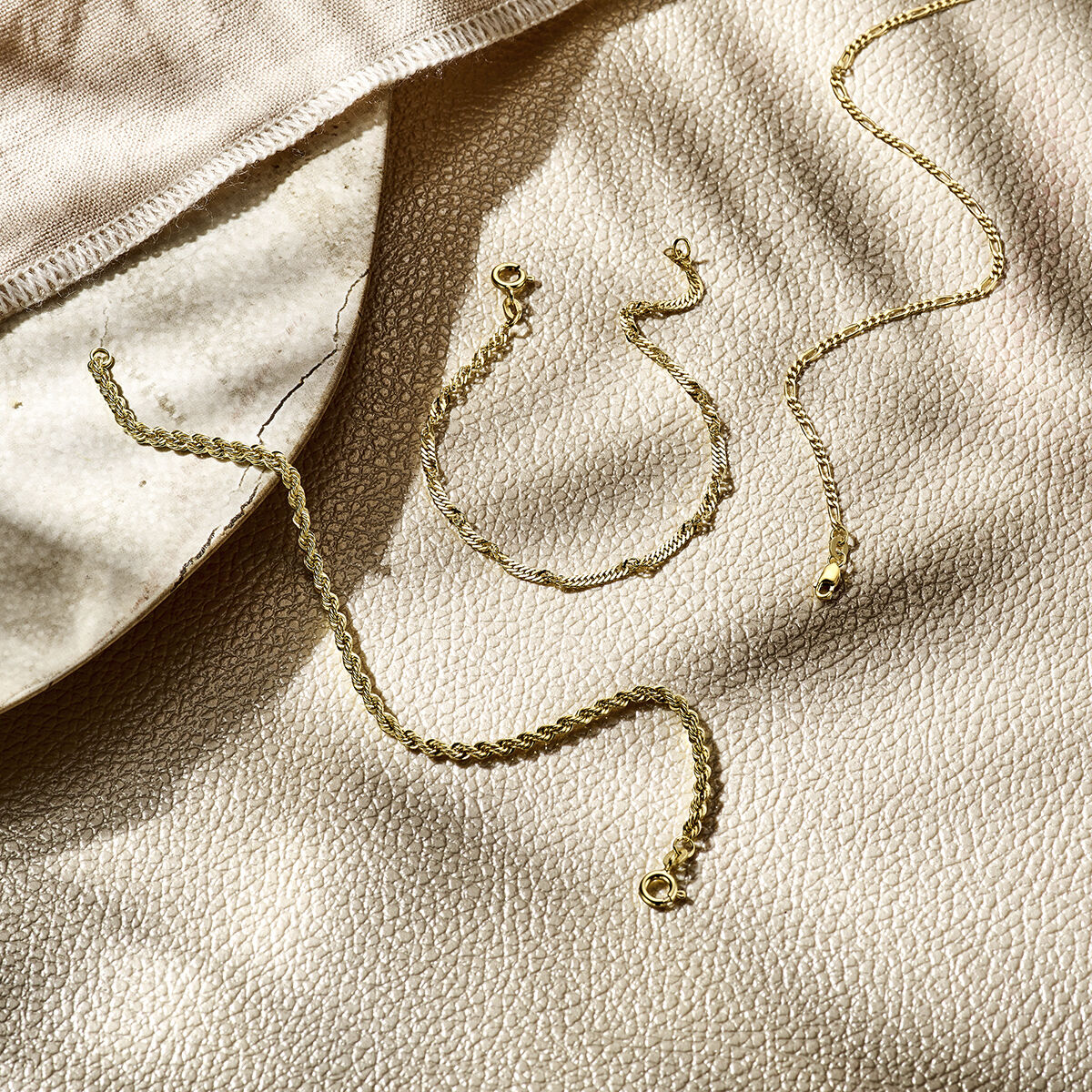 De armband figaro 1,8 mm 14K geelgoud van Van der Kooij Collectie wordt getoond op een crèmekleurig oppervlak met textuur, naast een beige doek en een keramische schaal, alles verlicht door zacht natuurlijk zonlicht.