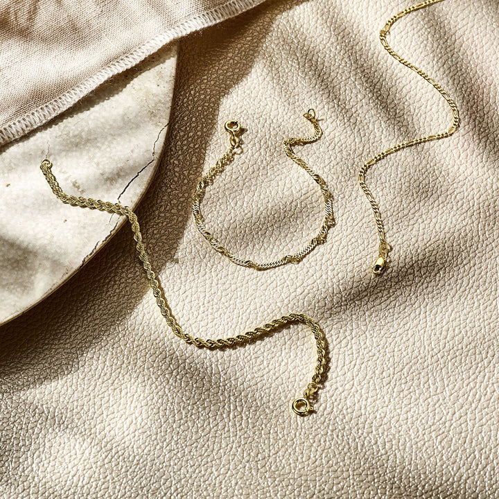 Drie delicate 14K geelgouden sieraden, waaronder de Van der Kooij Collectie armband figaro 1,8 mm, liggen op een beige oppervlak met zachte schaduwen en een stukje lichte stof zichtbaar in de hoek.