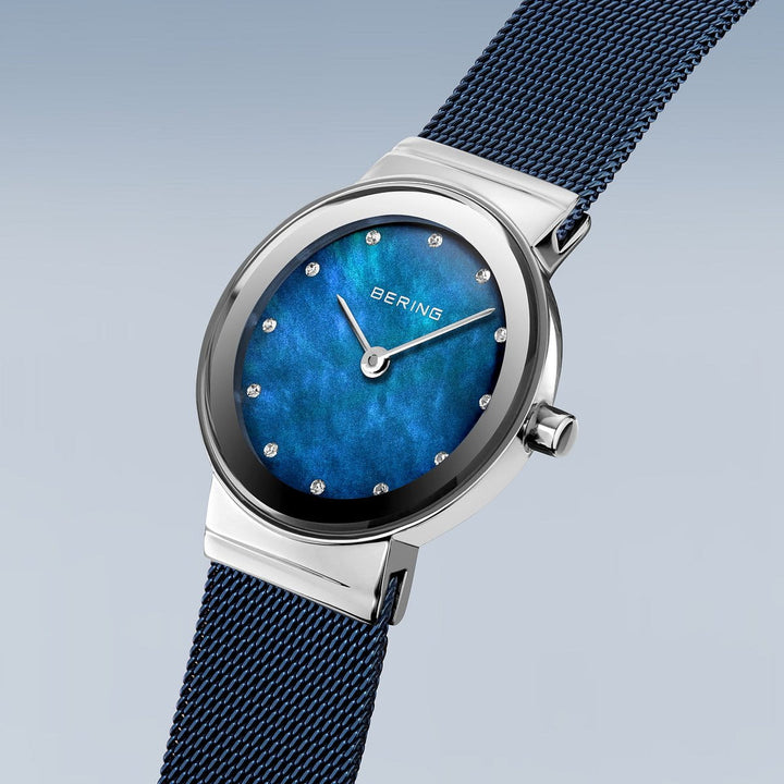 Het Bering 10126-307 dameshorloge heeft een blauwe parelmoer wijzerplaat, zilveren uurmarkeringen en wijzers. Dit quartzhorloge heeft een roestvrijstalen kast, blauwe meshband en saffierglas, afgezet tegen een lichtblauwe naar grijze achtergrond met kleurverloop.