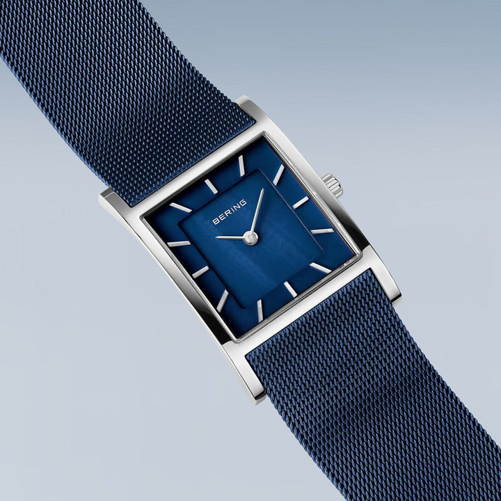 Het Bering dameshorloge 10426-307-S heeft een rechthoekig ontwerp met een blauwe wijzerplaat en meshband. Het bevat zilveren uurmarkeringen, wijzers en merknaam, afgezet tegen een lichtblauwgrijze achtergrond met kleurverloop. De zilveren kast wordt beschermd door duurzaam saffierglas en wordt aangestuurd door quartz uurwerk.