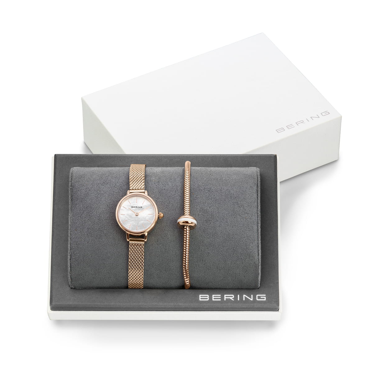 Reloj con esfera blanca y correa de malla rosa, acompañado de pulsera delgada dorada.