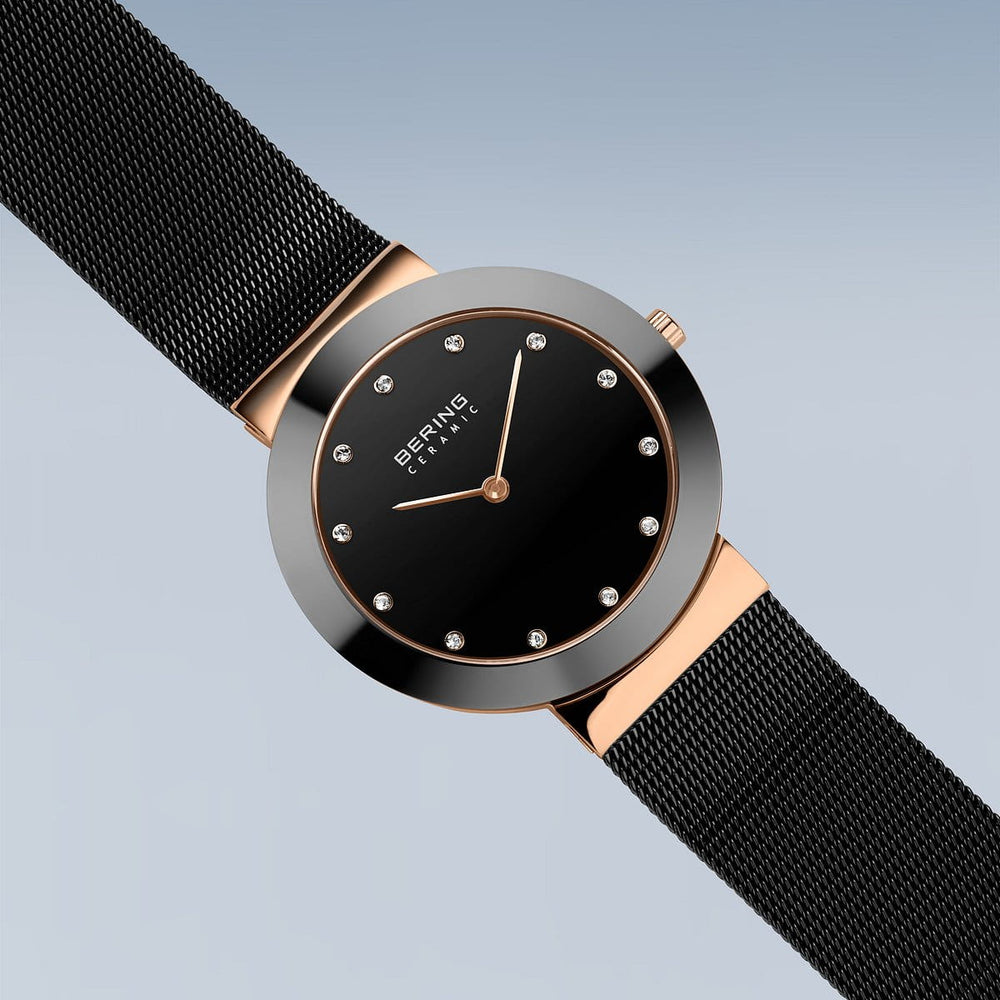 Het Bering 11435-166 dameshorloge heeft een zwarte meshband en gouden lunette. Het minimalistische quartz-ontwerp heeft een strakke zwarte wijzerplaat met uurmarkeringen, de merknaam in het midden en duurzaam saffierglas tegen een lichtblauwe achtergrond met kleurverloop.