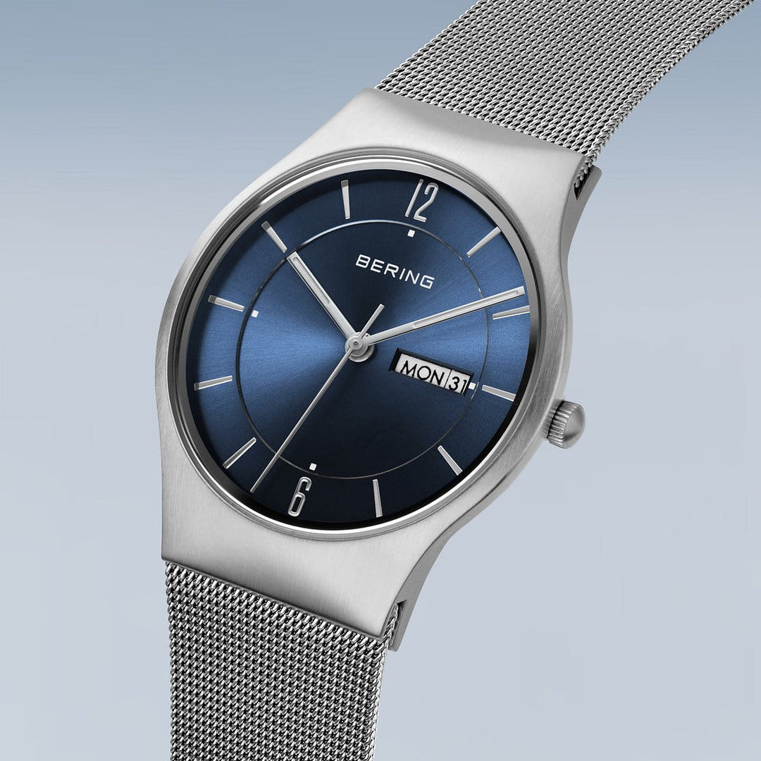 Hier ziet u een Bering 11938-003DD horloge met een zilveren roestvrijstalen kast, blauwe wijzerplaat, quartz uurwerk en een datumvenster met de tekst MON 31. Het minimalistische ontwerp bestaat uit saffierglas en een strakke meshband tegen een achtergrond met een lichtblauwe tot grijze kleurovergang.