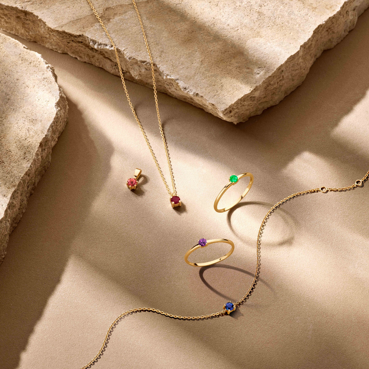 Van der Kooij Collectie’s 14K geelgouden ring met geboortesteen smaragd (mei), samen met twee kettingen en oorbellen, liggen op een beige steenoppervlak waar zachte zonlicht schaduwen werpt.