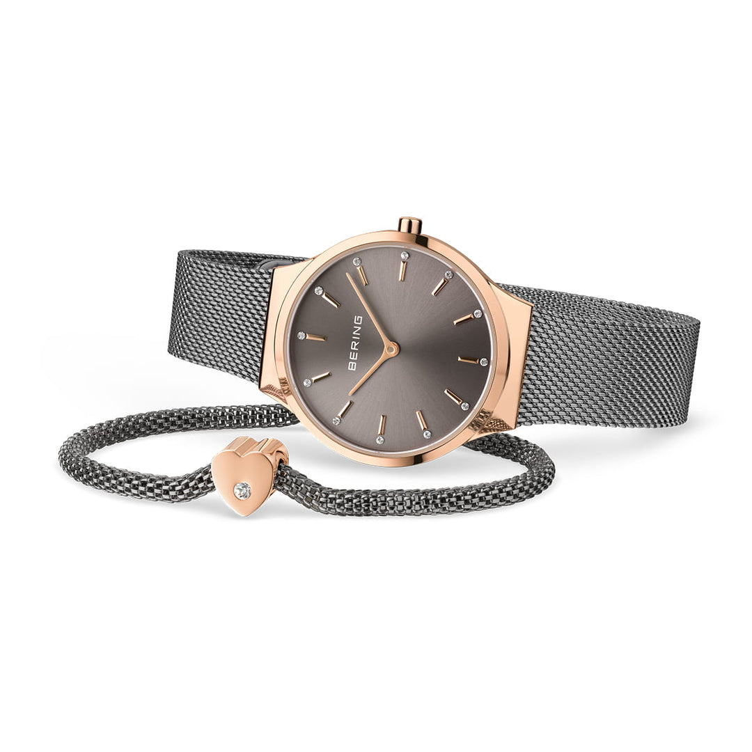 Een roségouden en grijs Bering horloge (12131-369-GWP) met een strak quartz uurwerk ligt op zijn kant. Ernaast, een bijpassende grijze Milanese band armband met een kleine bedel versierd met een kristal, maakt dit moeiteloos elegante duo compleet.