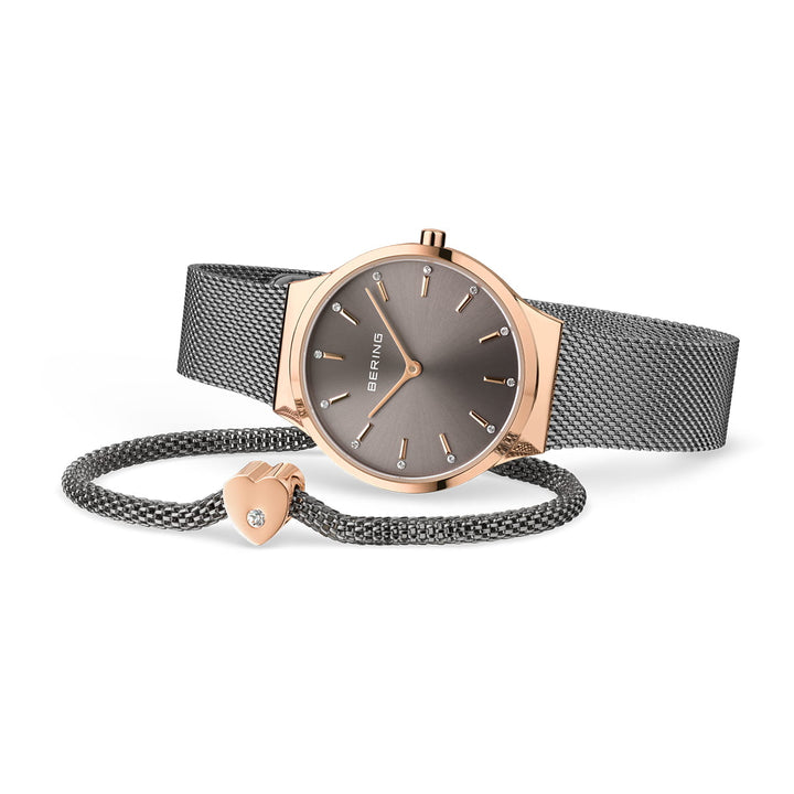 Een roségouden en grijs Bering horloge (12131-369-GWP) met een strak quartz uurwerk ligt op zijn kant. Ernaast, een bijpassende grijze Milanese band armband met een kleine bedel versierd met een kristal, maakt dit moeiteloos elegante duo compleet.