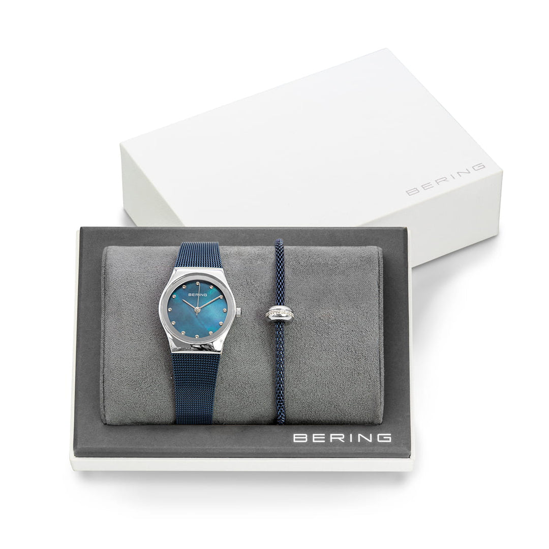 Het Bering 12927-307-GWP dameshorloge met Milanees bandje is een zilveren horloge met een blauwe wijzerplaat en band, gecombineerd met een bijpassende blauwe armband, elegant gepresenteerd op een getextureerd dienblad en genesteld in een elegante witte Bering doos.