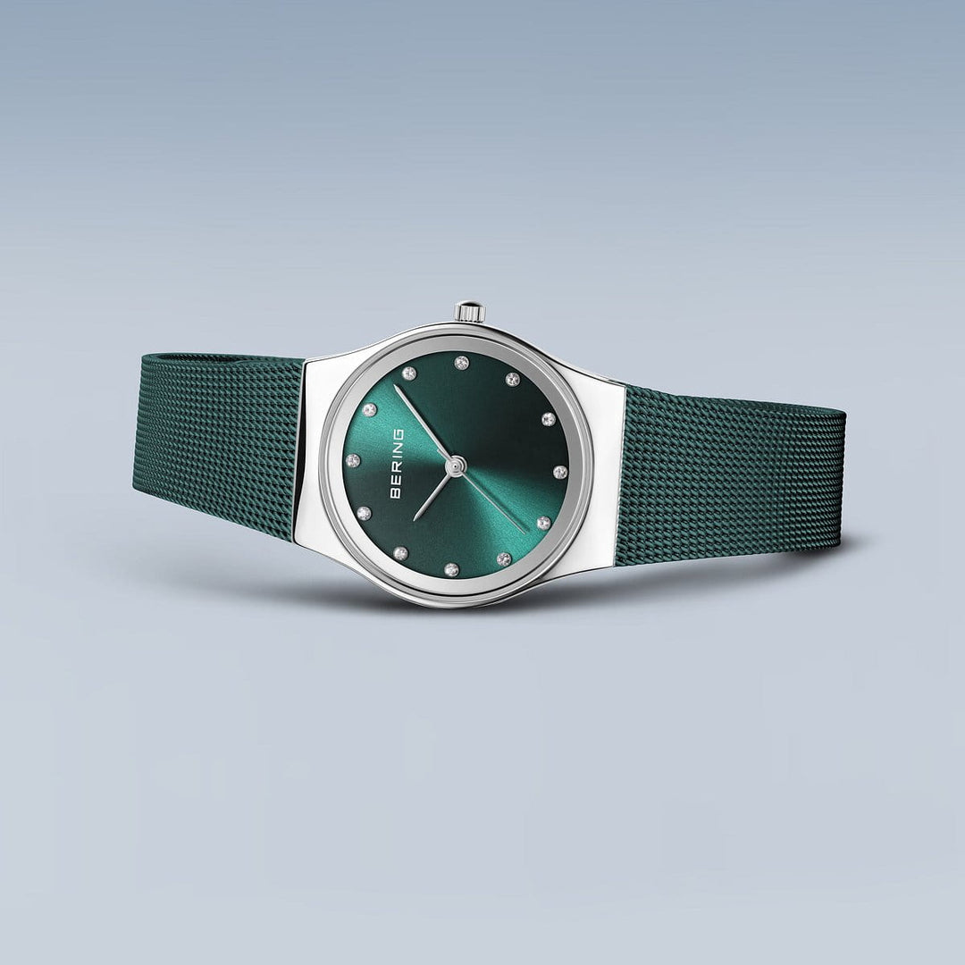 Het Bering Dameshorloge, model 12927-808, heeft een groene wijzerplaat met puntmarkeringen en het BERING-logo. De zilveren kast is gecombineerd met een getextureerd Milanees bandje, allemaal op een grijze achtergrond, wat de tijdloze Quartz-uurwerkprecisie belichaamt.