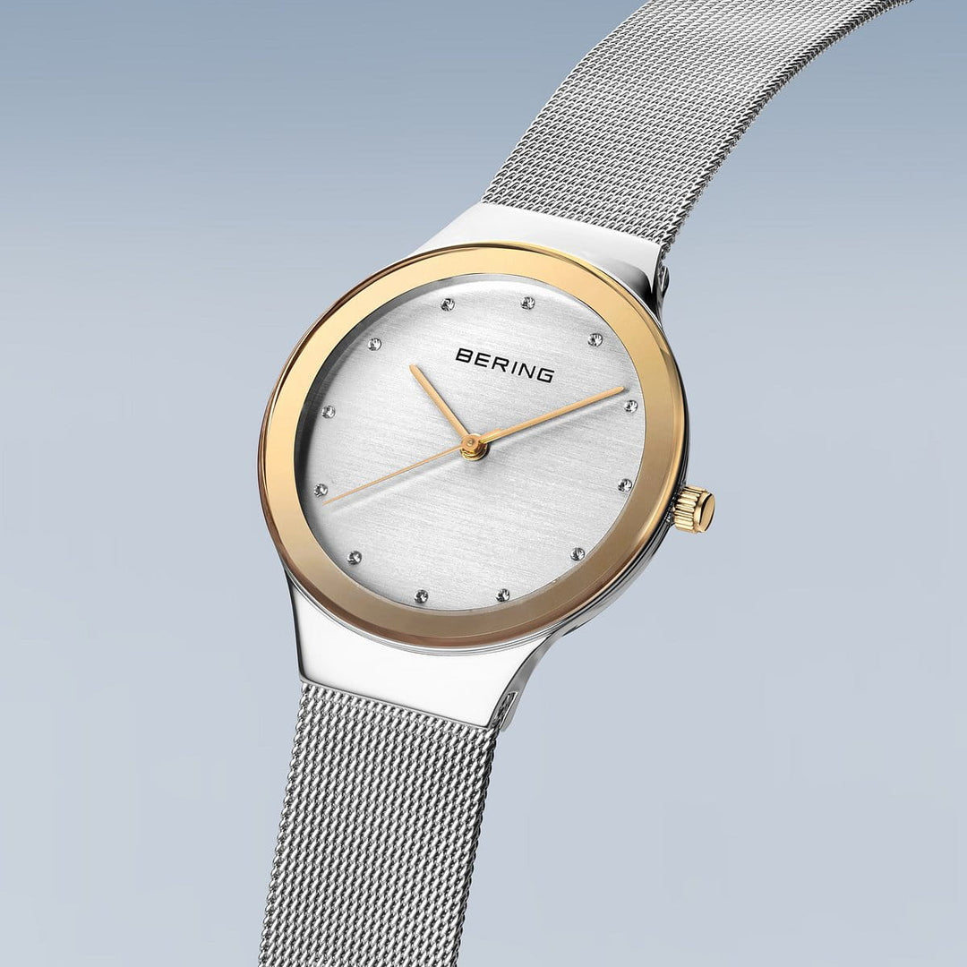De Bering 12934-010 is een strak dameshorloge met een tijdloos design, voorzien van een roestvrijstalen kast en zilveren mesh band. Het heeft een witte wijzerplaat met gouden lunette, zilveren en uurmarkeringen. De naam Bering valt op op de lichtblauwe gradient achtergrond.