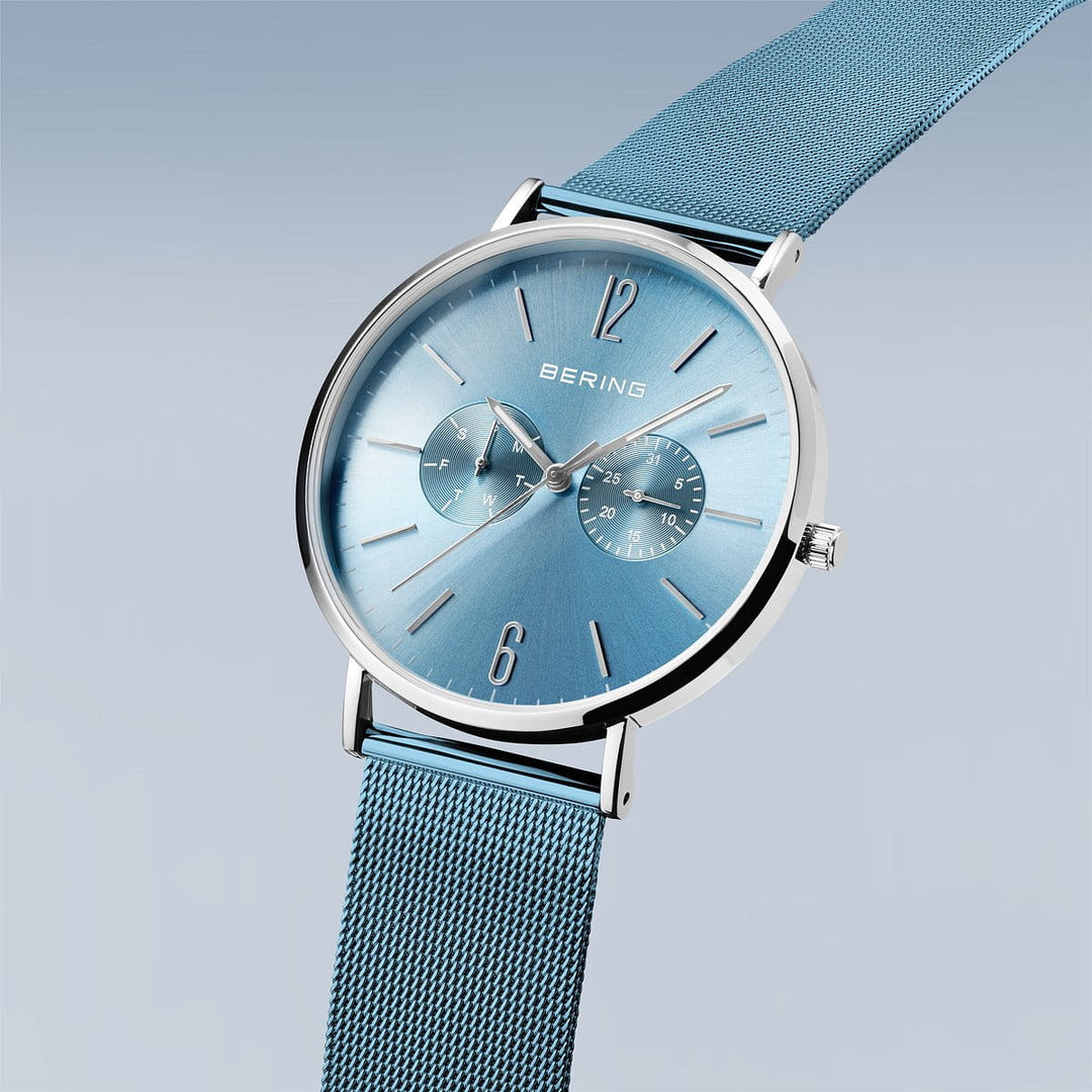 Product 14240-809 van Bering is een stijlvol herenhorloge met een blauwe sunray wijzerplaat, twee subdials, roestvrijstalen kast, blauwe mesh band en quartz-uurwerk tegen een lichtblauwe achtergrond.