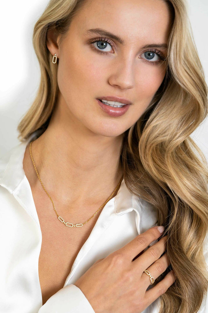 Een vrouw met lang blond haar draagt een witte zijden blouse, Zinzi Oorbellen Gold Plated Zio2398 Y oorbellen, een delicate gouden ketting en een gouden ring. Ze kijkt met een lichte glimlach naar de camera en raakt haar kraag aan.