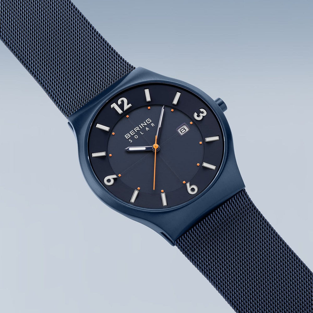 Het Bering-model 14440-393 wordt schuin weergegeven en heeft een blauw zonne-uurwerk en een mesh-band. Het horloge heeft een zwarte wijzerplaat met witte markeringen, een secondewijzer met oranje punt en een klein datumvenster onder duurzaam saffierglas op een achtergrond die van licht naar donkerblauw verloopt.