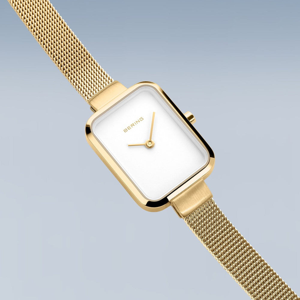 Het Bering dameshorloge 14520-334 heeft een roestvrijstalen kast, rechthoekige witte wijzerplaat, minimalistisch design, gouden wijzers en een goudkleurige mesh band op een lichtblauwe achtergrond.