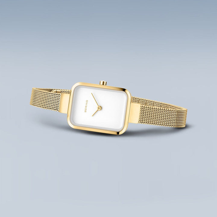 Het Bering horloge 14520-334 wordt getoond tegen een lichtblauwe achtergrond. Dit goudkleurige model heeft een vierkante witte wijzerplaat, minimalistisch design, mesh metalen band en gouden wijzers en roestvrijstalen kast.