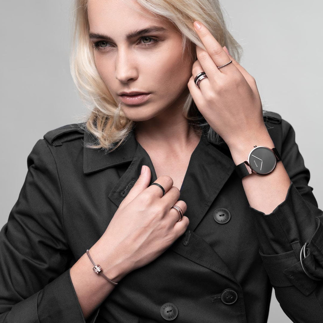 Een blondine in een zwarte trenchcoat toont ringen, een armband en het Bering horloge model 14539-166 met een roestvrijstalen kast op hun linker pols. Ze kijken naar links met een neutrale uitdrukking tegen een effen achtergrond.