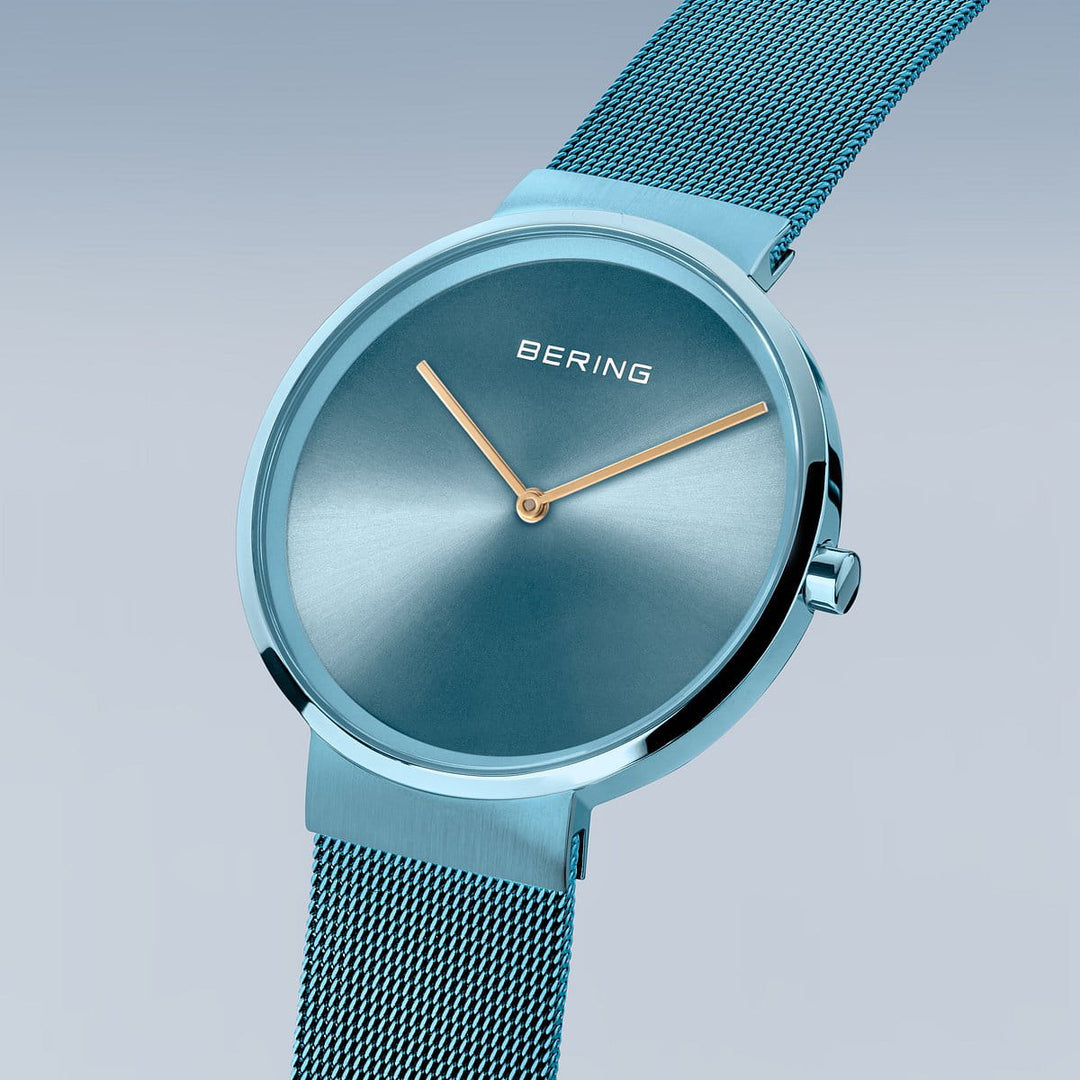 Product 14539-388 van Bering is een strak dameshorloge met een turquoise metallic finish, gouden uur- en minutenwijzers, quartz-uurwerk, saffierglas en een fijn mesh bandje. Het minimalistische design heeft een ronde wijzerplaat en de merknaam siert de wijzerplaat tegen een zachte gradiënt achtergrond.