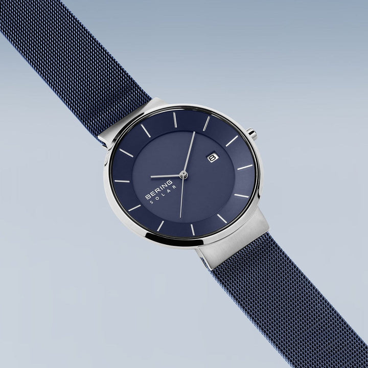 De Bering 14639-307 heeft een strak design met een blauwe mesh band en ronde blauwe wijzerplaat, minimalistische zilveren uurmarkeringen, wijzers en een datumvenster op 3 uur beschermd door duurzaam saffierglas. Het wordt getoond tegen een lichtblauwe achtergrond.