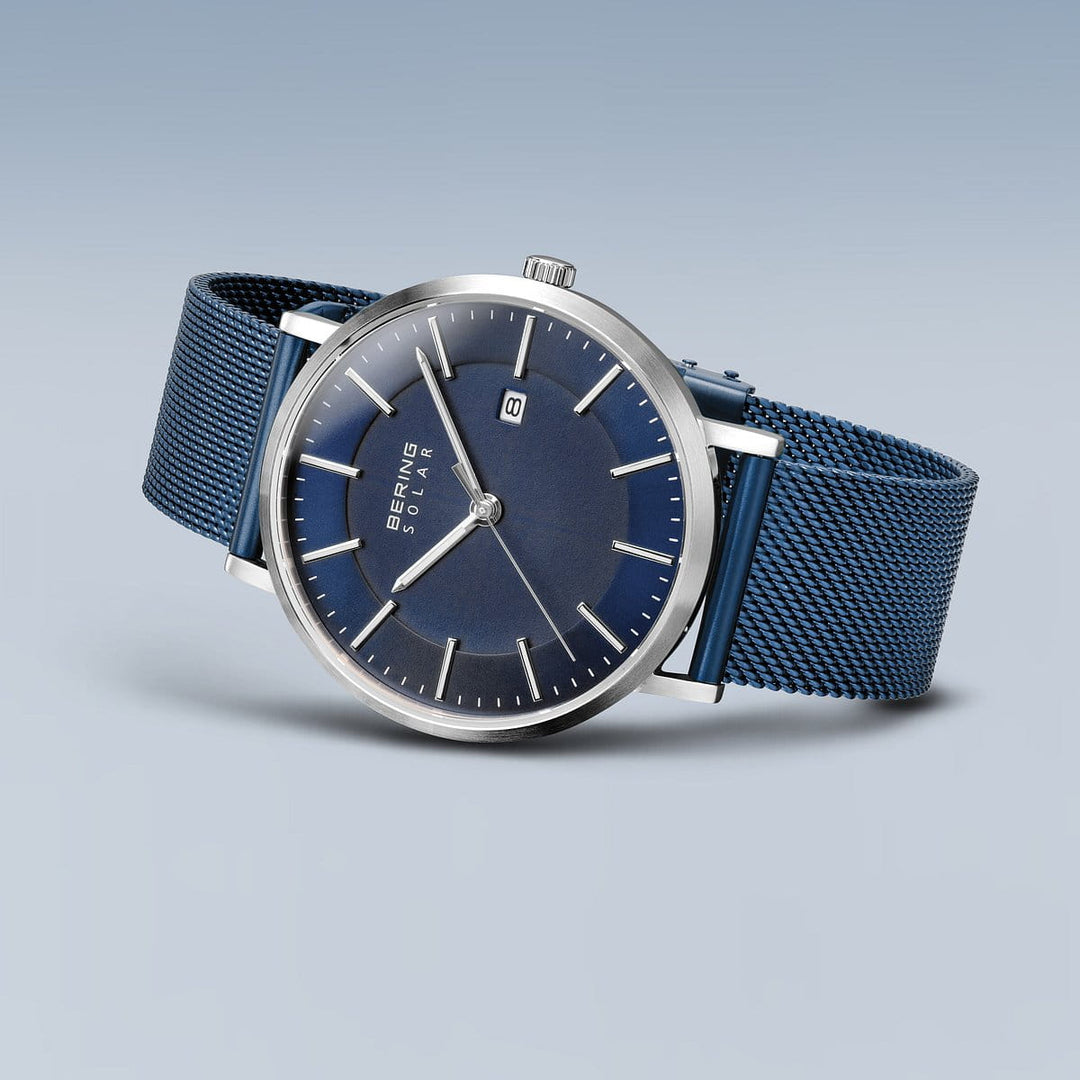 Ontdek het elegante Bering horloge (15439-307) met een blauwe mesh band, saffierglas, zilveren uurmarkeringen en wijzers, plus een datumvenster. Op zonne-energie in een metalen behuizing, het heeft een strak design tegen een lichtblauwe achtergrond.