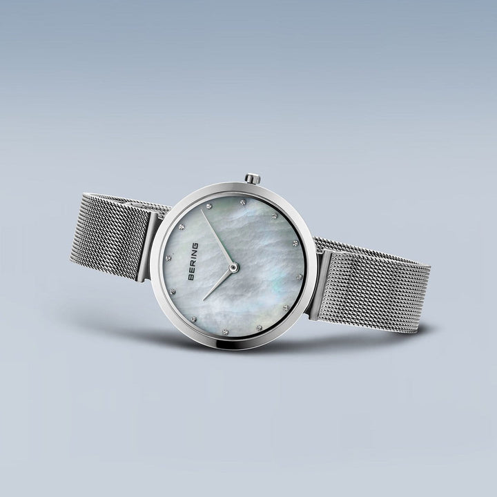 Het Bering 18132-004 dameshorloge heeft een zilveren meshband en een minimalistisch design op een lichtblauwe achtergrond. De roestvrijstalen kast omringt een ronde parelmoeren wijzerplaat met saffierglas en eenvoudige uurmarkeringen.