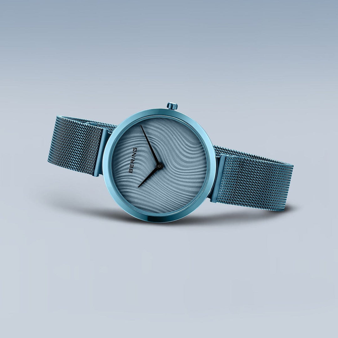 De Bering dames horloge blauwe wijzerplaat - 18132-Charity2 heeft een minimalistische blauwe wijzerplaat, mesh horlogeband, eenvoudige zwarte wijzers en quartz uurwerk van Bering, weergegeven op een lichtblauwe gradiënt achtergrond.