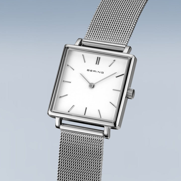 Het Bering dameshorloge, model 18226-004, heeft een vierkant zilveren polshorloge met een witte wijzerplaat, minimalistische uurmarkeringen en een dunne meshband. Het heeft quartz uurwerk en saffierglas voor duurzaamheid. Het strakke ontwerp is afgezet tegen een lichtblauwe gradiëntachtergrond.
