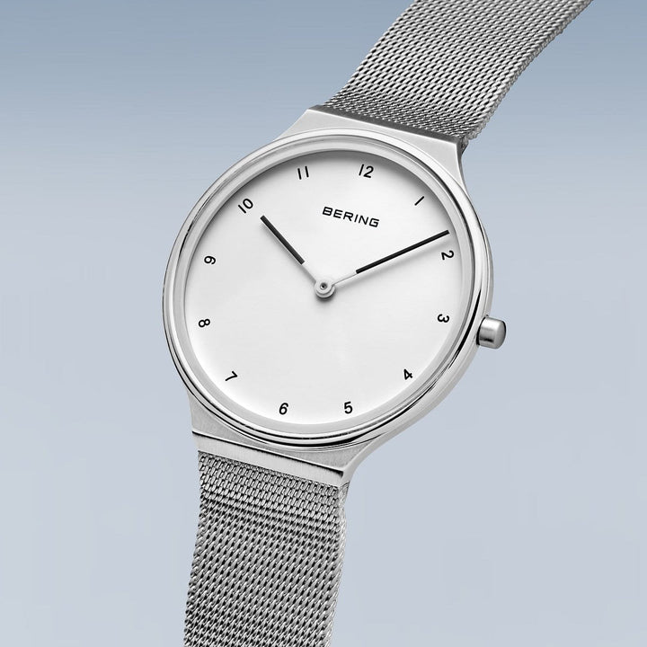 Een strak Bering dameshorloge (18434-004) met een minimalistisch design, voorzien van een mesh band en quartz uurwerk. Het heeft een witte wijzerplaat, zwarte uurmarkeringen, twee dunne wijzers, duurzame saffierglas bescherming en BERING weergegeven onder 12 uur tegen een blauwe gradiënt.