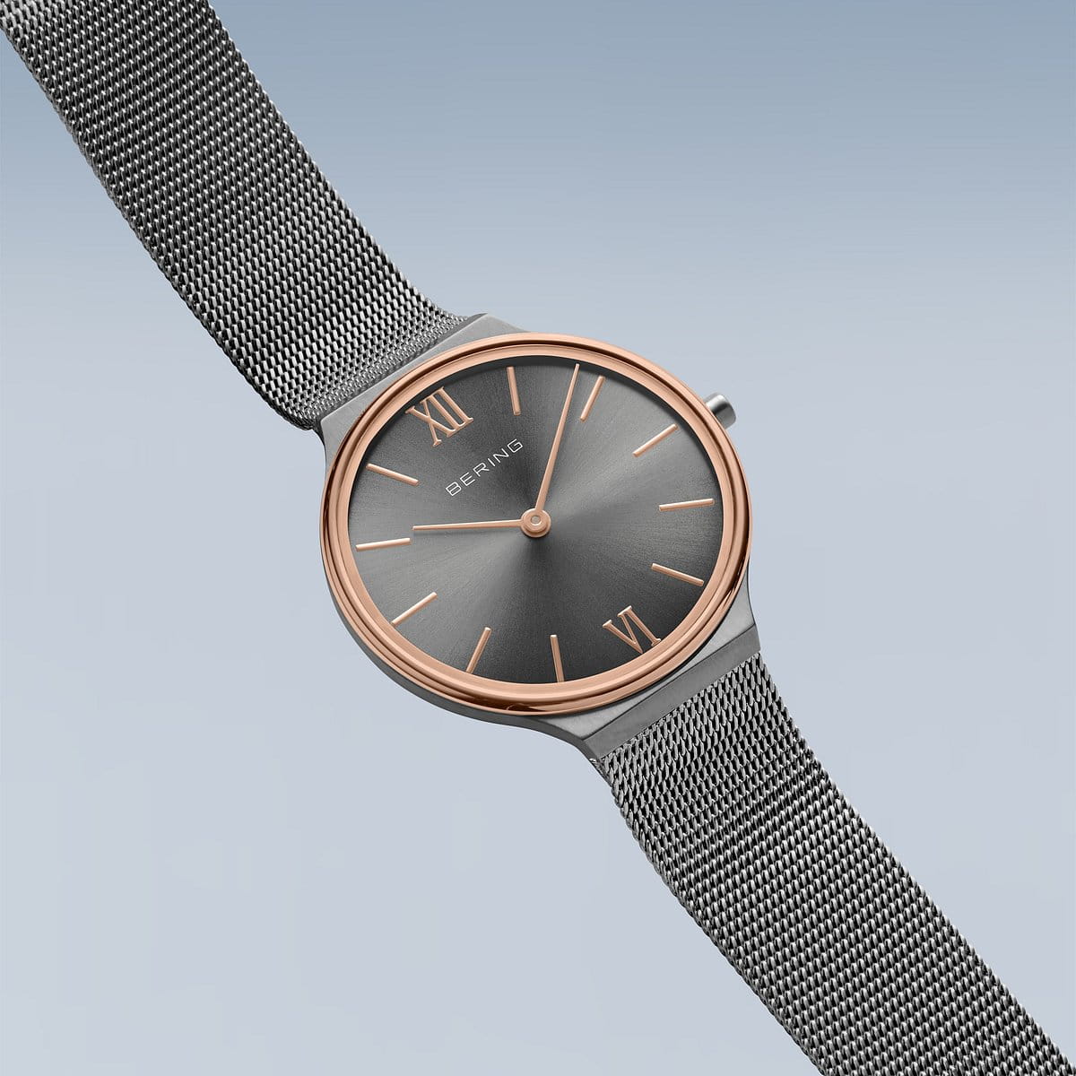 Het Bering dameshorloge 18434-369 heeft een strak, minimalistisch design met een metalen mesh band, een donkergrijze wijzerplaat met een roségouden accent, een quartz uurwerk beschermd door saffierglas, Romeinse cijfers op 12 en 6 uur en eenvoudige uurmarkeringen op een zachtblauwe achtergrond.