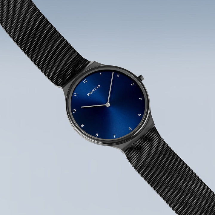 Het Bering dameshorloge (18440-227) heeft een strakke blauwe wijzerplaat met minimalistische uurmarkeringen en zilveren wijzers, gevat in een roestvrijstalen kast. Het is gecombineerd met een zwarte Milanees band en elegant geplaatst tegen een lichtblauwe gradiëntachtergrond.