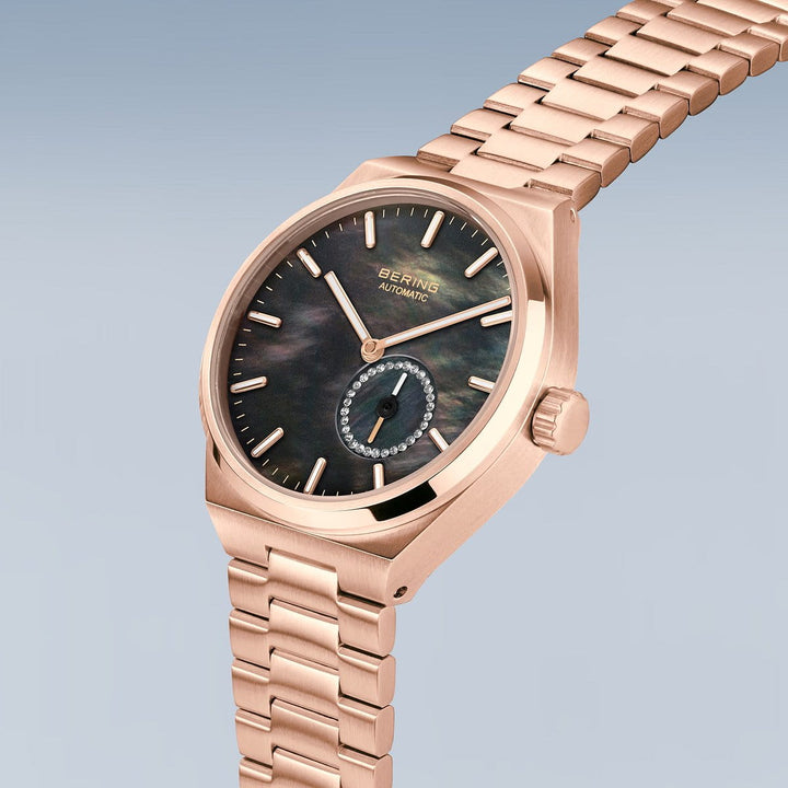 De Bering dames horloge zwart dial - 19435-762 heeft een roségouden polshorloge met een metalen schakelband en roestvrijstalen kast. De donkere wijzerplaat is versierd met roségouden uurmarkeringen, wijzers, een met kristal geaccentueerde subdial en de kroon rust sierlijk op de rechterkant.