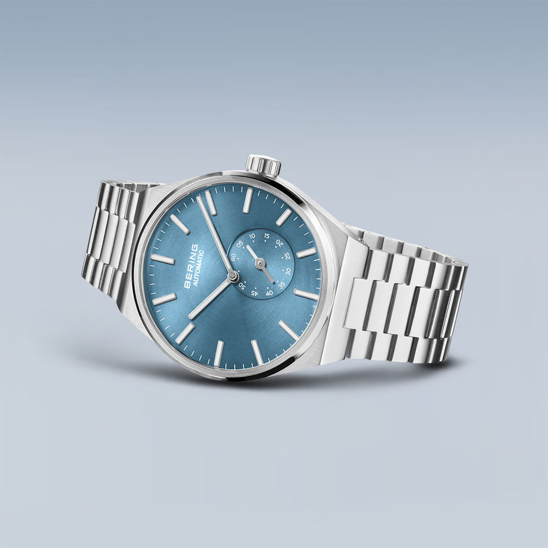 Het Bering herenhorloge model 19441-Charity heeft een blauwe wijzerplaat met zilveren uurmarkeringen en saffierglas, een secundaire wijzerplaat en een metalen schakelband op een lichtblauwe achtergrond met kleurverloop.