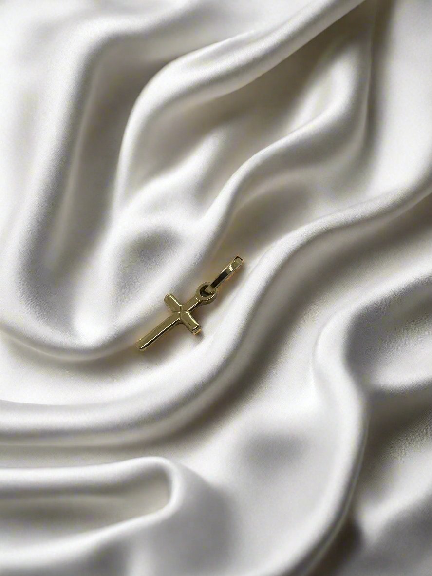 Colgante de cruz en oro amarillo de 14K, con un peso de 0.8 gramos, sobre fondo de satén blanco.
