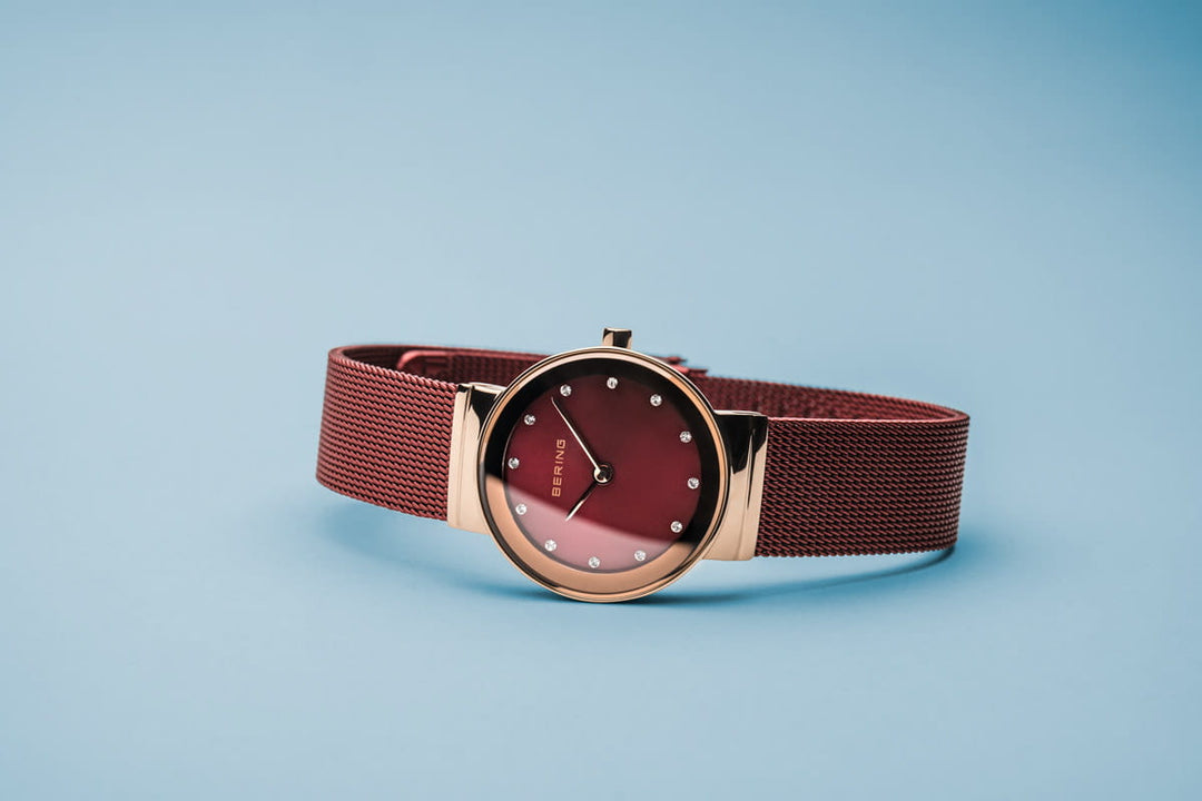 De Bering 10126-363 is een minimalistisch dameshorloge met een roségouden kast en een dieprode wijzerplaat met stipmarkeringen. De rode meshband complementeert het Quartz uurwerk, alles tegen een lichtblauwe achtergrond voor een elegante look.