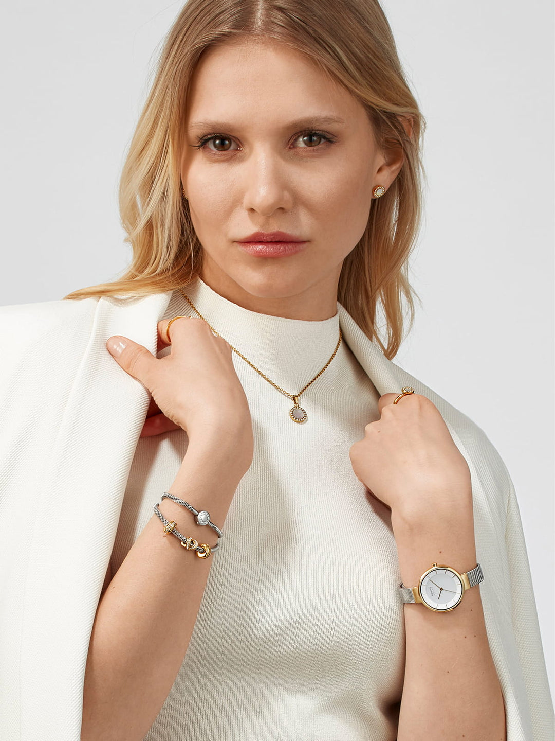 Een langharige blondine draagt een witte jas over een licht topje en draagt daarbij een gouden ketting, armbanden en oorstekers. Daarnaast draagt ze het Bering dames horloge witte dialekt - 14631-024 met quartz-uurwerk en saffierglas. Ze poseert tegen een effen achtergrond.