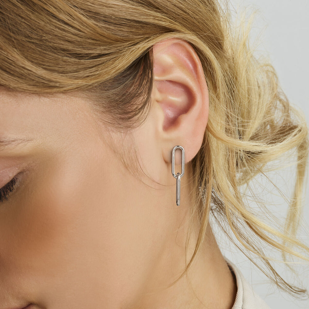 Close-up van het oor van een vrouw met Van der Kooij Collectie oorhangers paperclip zilver gerhodineerd. De langwerpige, geometrische 925 zilveren oorbellen steken af tegen haar lichte huid en blonde haar in losse golven.