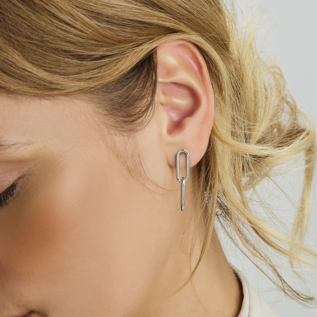 Close-up van het oor van een vrouw met Van der Kooij Collectie oorhangers paperclip zilver gerhodineerd. De langwerpige, geometrische 925 zilveren oorbellen steken af tegen haar lichte huid en blonde haar in losse golven.