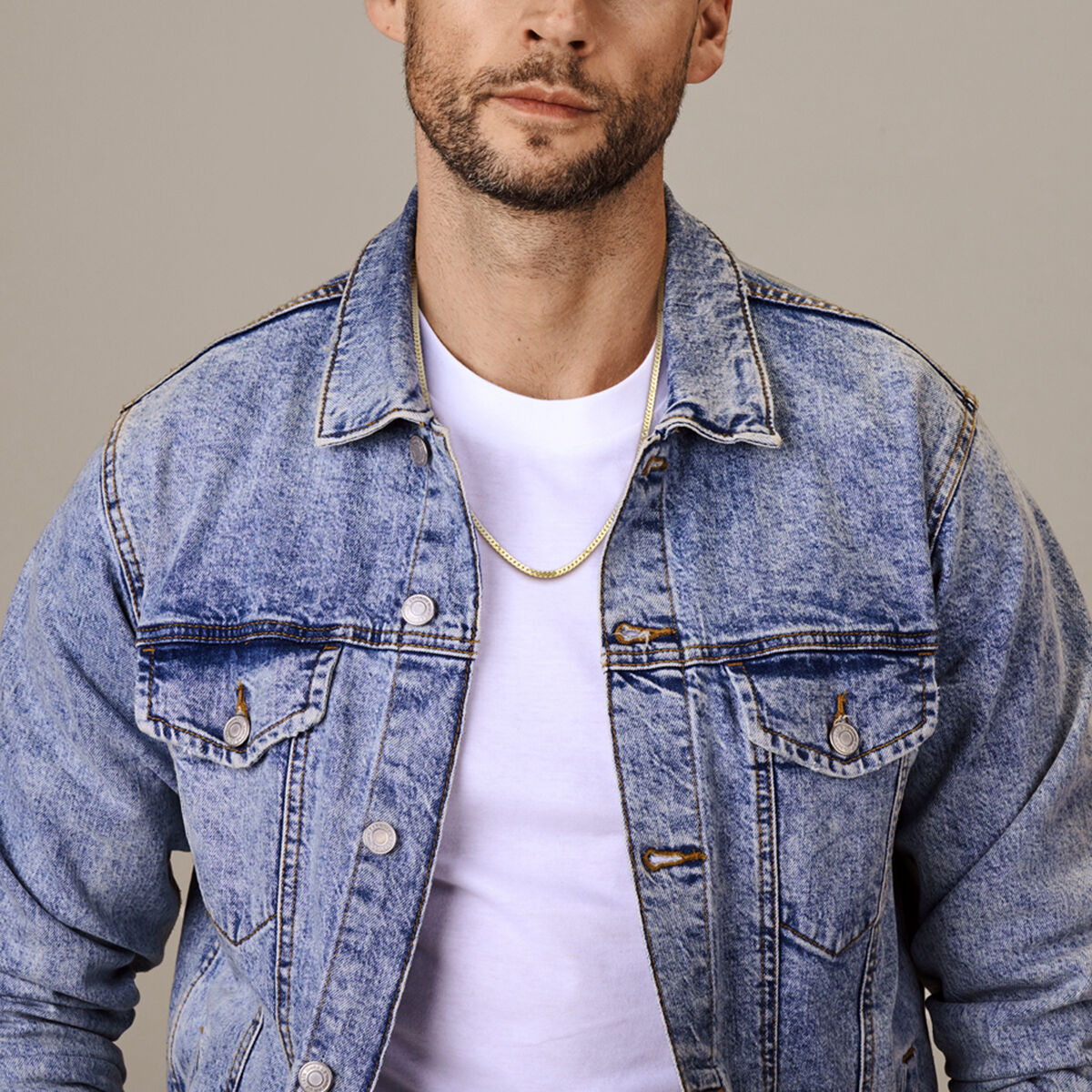 Een man draagt een lichtblauw spijkerjack over een wit T-shirt en de Van der Kooij Collectie Gouden ketting heren - gourmette 4-zijdes geslepen 2,7 mm massief 14K, afgebeeld van schouders tot middenborst tegen een neutrale achtergrond.