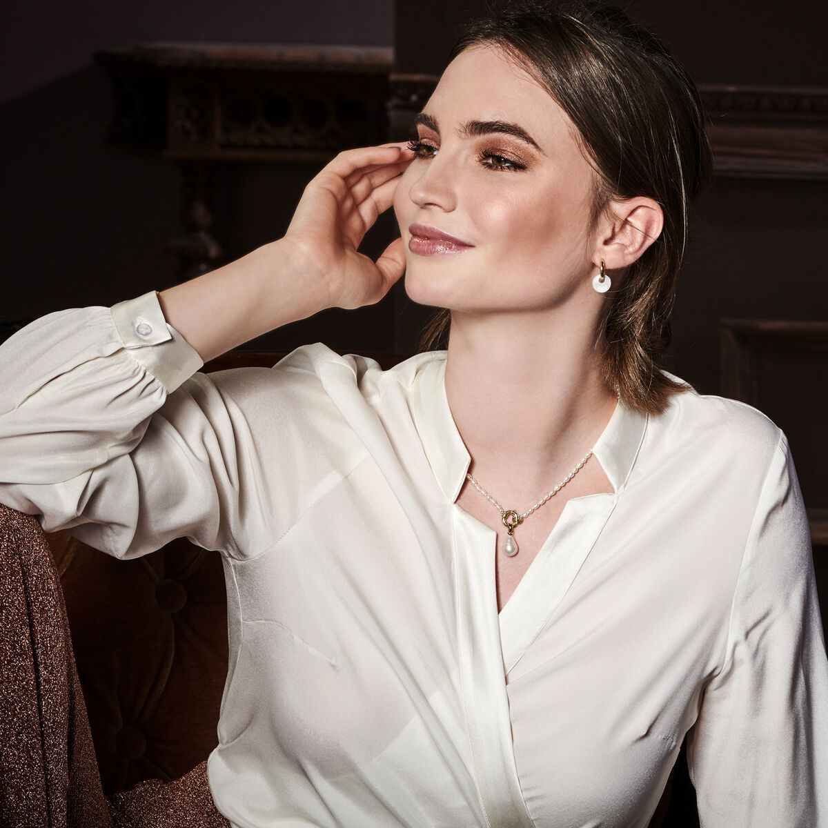 Een vrouw met een lichte huid en bruin haar, draagt een witte blouse en het Van der Kooij Collectie hanger parel 14K geelgoud collier, glimlacht zittend op een bruine stoel in een zacht verlichte kamer.