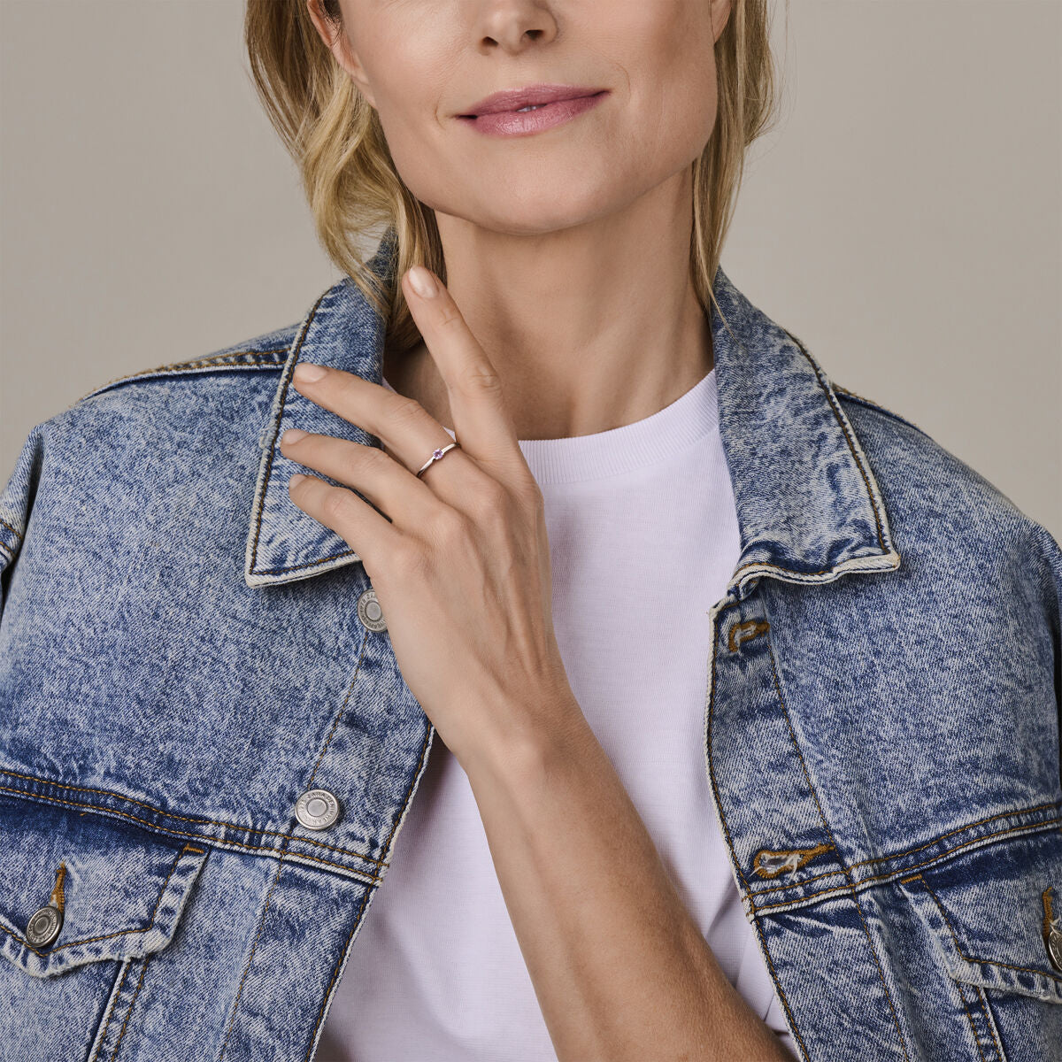 Een vrouw in een licht spijkerjasje en wit shirt betast haar gezicht en toont de Van der Kooij Collectie ring met geboortesteen licht amethist juni zilver gerhodineerd aan haar linkerhand.