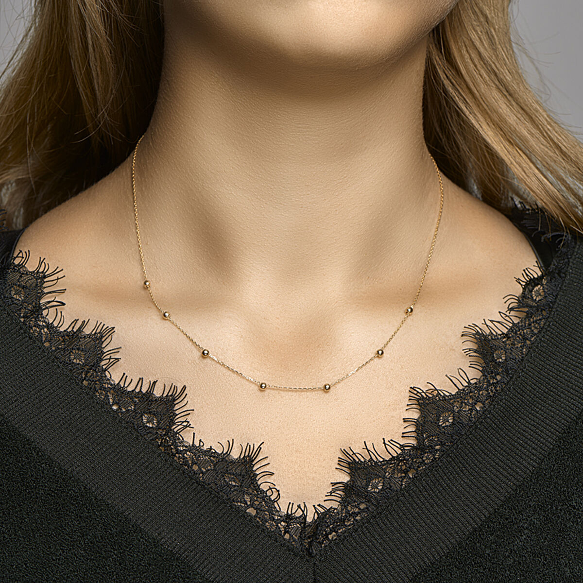 Een vrouw draagt een Van der Kooij Collectie collier anker en bolletjes 1,0 mm 42-44-46 cm 14K geelgoud, de delicate gouden kralen verspreid langs de ketting, gecombineerd met een zwart kanten topje. Alleen haar nek en bovenborst zijn zichtbaar.
