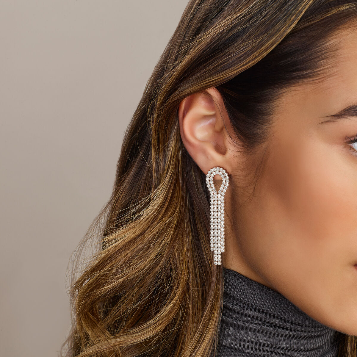Close-up van een vrouwenoor met Van der Kooij Collectie oorhangers zirkonia zilver gerhodineerd met meerdere strengen. Haar gladde bruine haar en geribbelde grijze top met hoge nek steken af tegen een neutrale achtergrond.