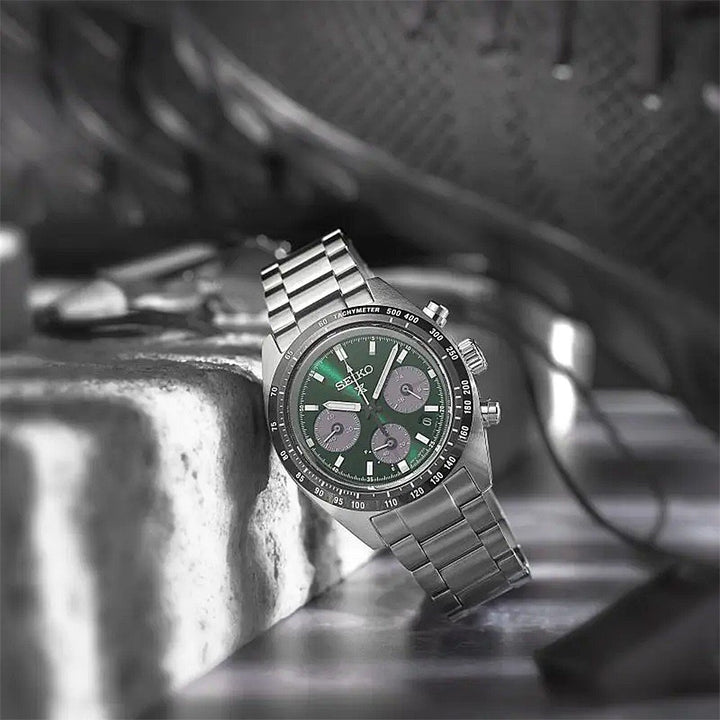 De Seiko Prospex SSC933P1, een zilveren herenhorloge met een groene wijzerplaat en drie sub-dials, wordt getoond op een metalen oppervlak. De contrasterende wijzers en markeringen van het horloge benadrukken de merknaam Seiko tegen de groene achtergrond, met achtergrondelementen die subtiel zijn vervaagd.