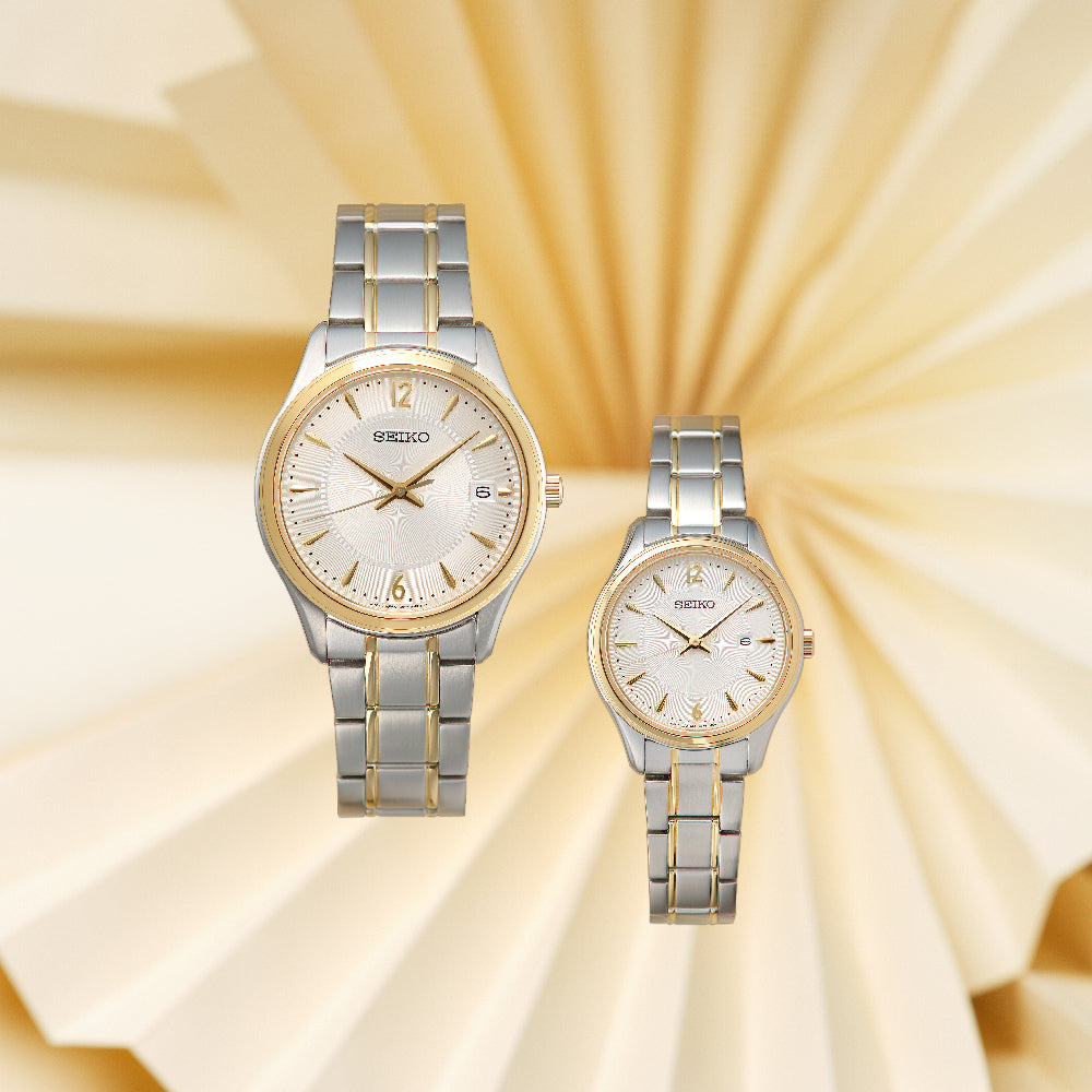 Twee zilveren en gouden horloges, waaronder een Seiko dameshorloge SUR474P1, rusten tegen een crèmekleurige geplooide papieren achtergrond. Het grotere horloge heeft een datumweergave en saffierglas; het kleinere horloge weerspiegelt het ontwerp zonder die functies. Beide hebben roestvrijstalen kasten voor extra elegantie.