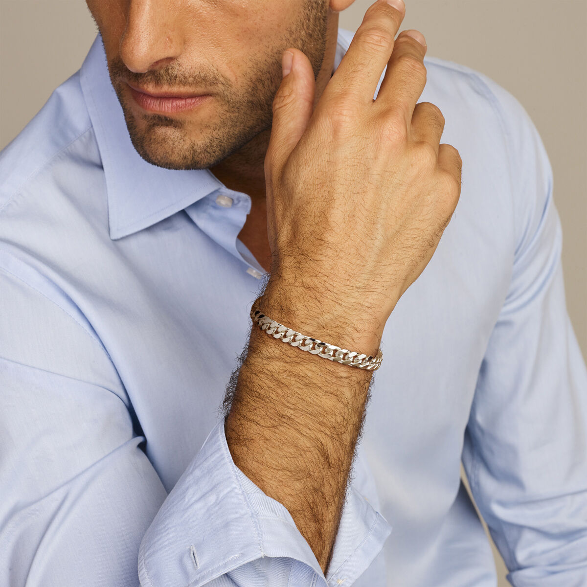 Een man met een lichtblauw overhemd draagt de Van der Kooij Collectie Zilveren armband heren gourmette 7,0 mm om zijn pols. Hij laat zijn hand rusten bij zijn kin met een neutrale uitdrukking die tijdloze elegantie uitstraalt.