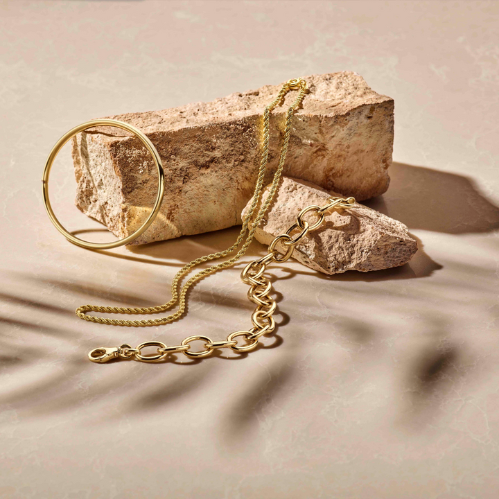 Een Van der Kooij Collectie armband scharnier massief ovale buis 4,0 mm Zilgoud (geelgoud met zilverkern) is gedrapeerd over twee beige steenblokken met textuur op een licht, marmerachtig oppervlak met subtiele schaduwen.