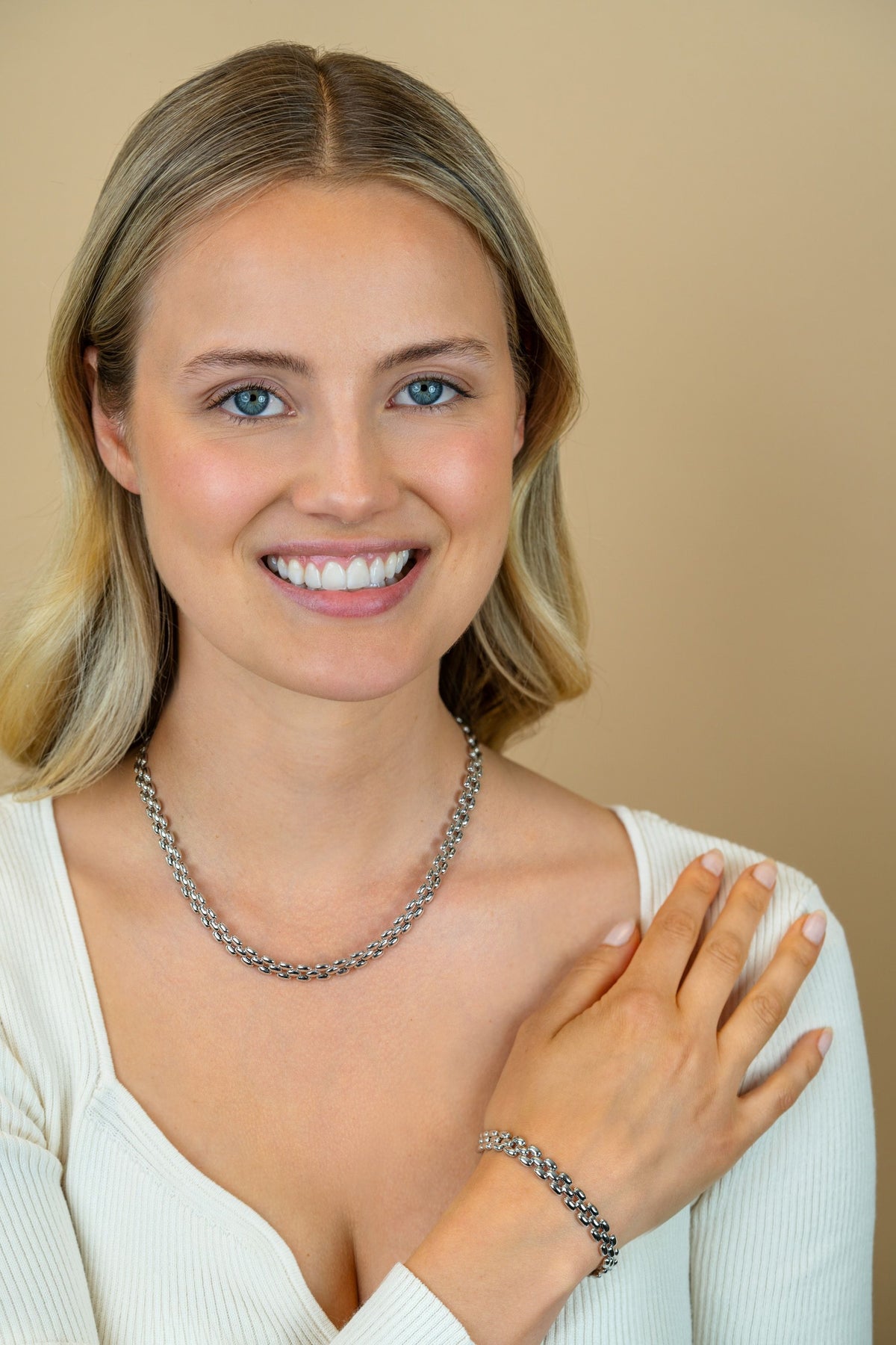 Een lachende blonde vrouw in een wit topje draagt de Zinzi Armband zilver ZIA2764 en een bijpassende zilveren ketting, met een hand op haar schouder tegen een beige achtergrond.