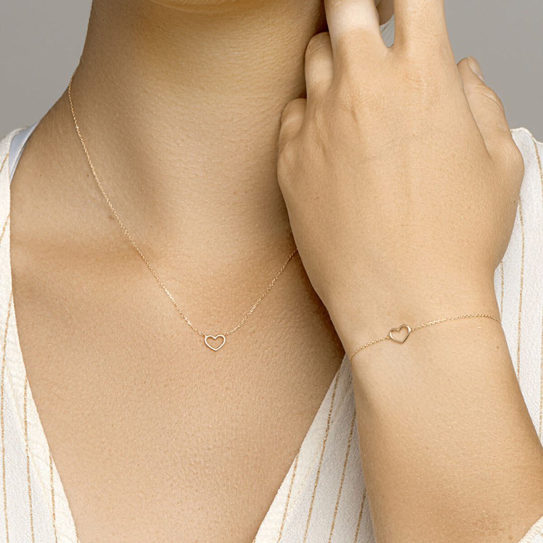 Een vrouw draagt de Van der Kooij Collectie Gouden armband dames hart 14K, een delicaat gouden armbandje met een open hart hanger, gecombineerd met een lichte huid en een witte V-hals - een ideale sieraad voor elke gelegenheid.