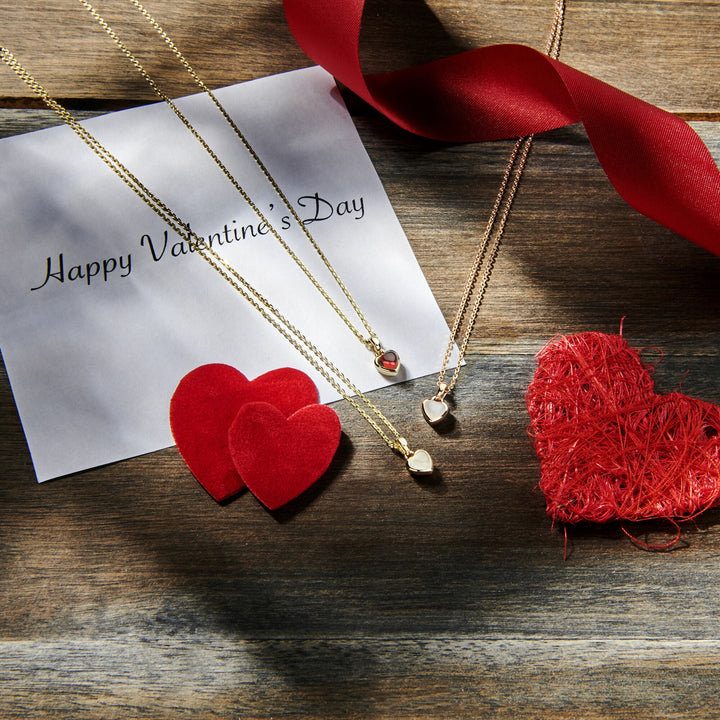 Twee hanger hart maansteen 14K roségouden kettingen van Van der Kooij Collectie, een kaart met de tekst Happy Valentines Day, rode stof en rieten harten, en een rood lint worden tentoongesteld op rustiek hout in fel zonlicht.