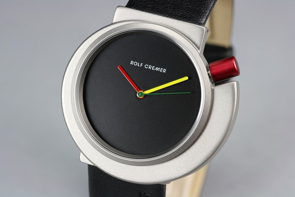 Het Rolf Cremer Horloge 492314 van Rolf Cremer is een modern unisex kwartshorloge met een zwarte wijzerplaat, minimalistisch ontwerp zonder cijfers, groene uurwijzer, gele minutenwijzer, rode secondewijzer, zilveren kast met rode kroon en zwarte leren band.