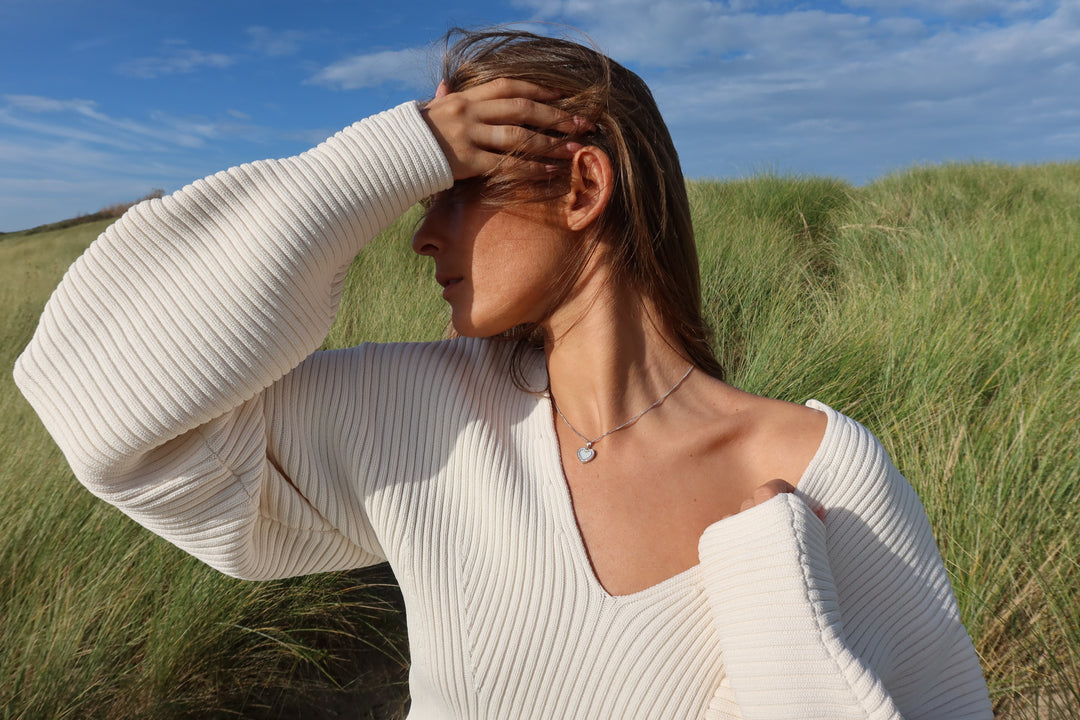 Een vrouw in een crèmekleurige trui staat in het hoge gras en raakt kalm haar voorhoofd aan, terwijl ze het elegante Demi | AsHanger massief | Zirkonia van Juwelier van der Kooij draagt onder een blauwe lucht, haar houding sereen en beschouwend.